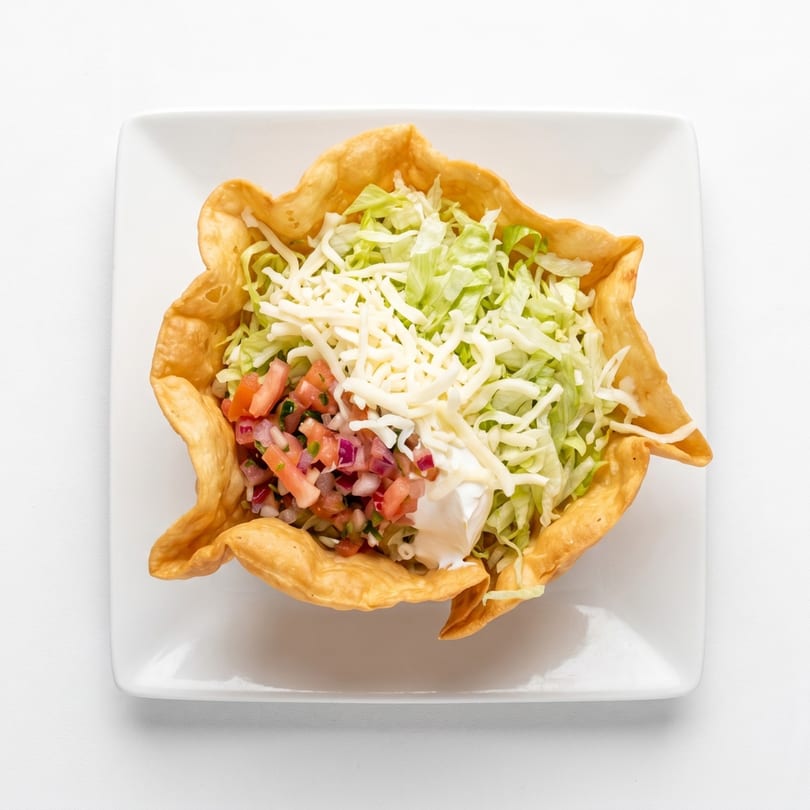 A taco salad in a crispy tortilla bowl with lettuce, cheese, pico de gallo, and sour cream.